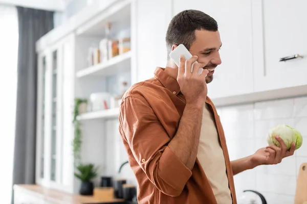 Hombre positivo hablando en el teléfono inteligente y la celebración de la col en la cocina - foto de stock