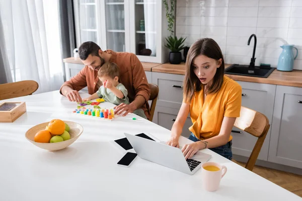 Mujer conmocionada usando portátil cerca de los dispositivos y la familia en casa - foto de stock