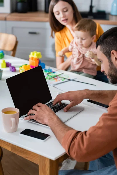 Freelancer usando laptop com tela em branco perto do copo e família turva em casa — Fotografia de Stock