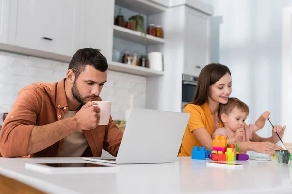 Young man holding cup while working on laptop near family and building blocks at home — Stock Photo