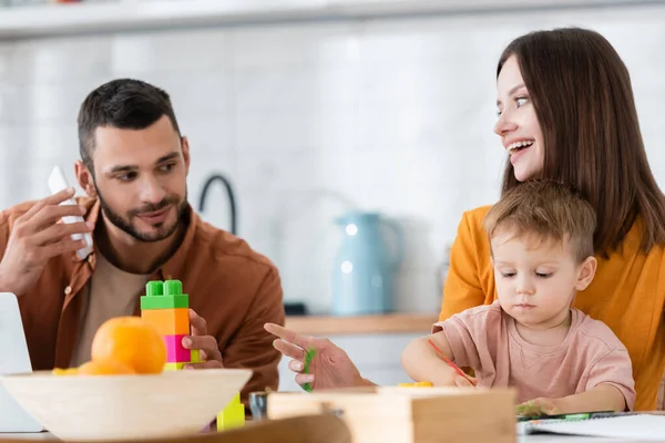 Positive Frau schaut Ehemann mit Smartphone an, während Sohn zu Hause zeichnet — Stockfoto