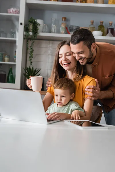 Alegre marido abrazando esposa con taza cerca de gadgets e hijo en casa - foto de stock