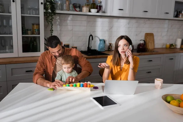 Freelancer jovem falando no smartphone perto de gadgets e jogo de família na cozinha — Fotografia de Stock