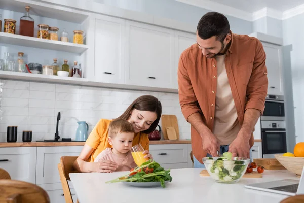 Mujer feliz sosteniendo jugo de naranja cerca de hijo y marido cortando verduras en la cocina - foto de stock
