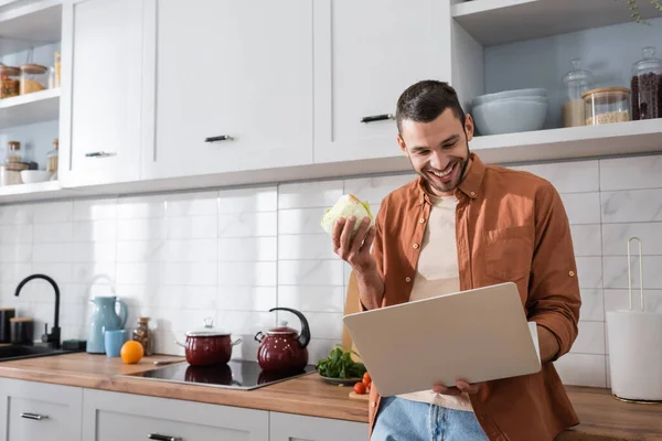 Un homme positif tenant du chou et regardant un ordinateur portable dans la cuisine — Photo de stock