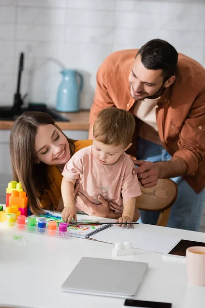 Sonriente familia mirando hijo cerca de bloques de construcción y cuaderno de bocetos en casa - foto de stock