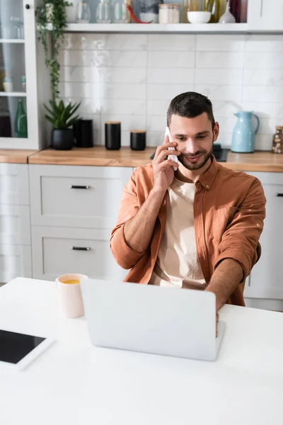 Young freelancer talking on smartphone and using laptop near cup of coffee in kitchen — Stock Photo