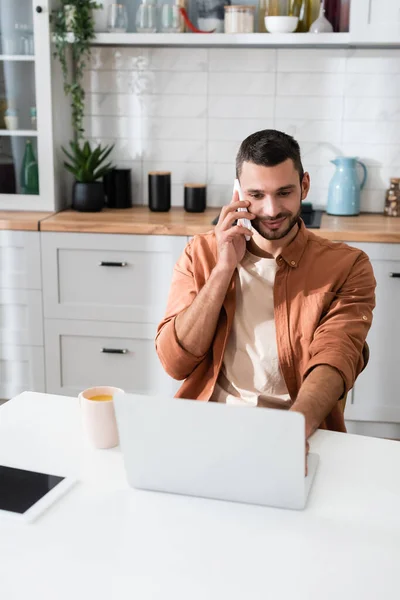 Jovem freelancer falando no smartphone e usando laptop perto de xícara de café na cozinha — Fotografia de Stock