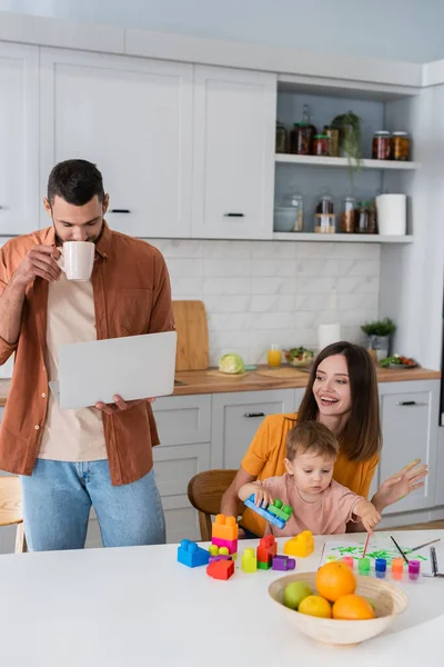 Madre e figlio giocare mattoni vicino marito con caffè e laptop in cucina — Foto stock