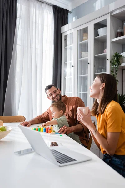 Cheerful woman holding cup near laptop and family playing game at home — Stock Photo