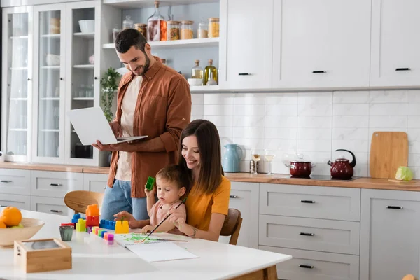 Mann benutzt Laptop und schaut sich Familienzeichnung in Küche an — Stockfoto