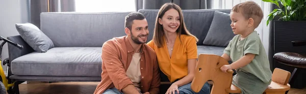 Lächelnde Eltern mit Blick auf Kleinkind auf Schaukelpferd zu Hause, Banner — Stockfoto