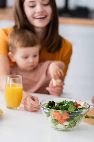 Ensalada fresca cerca del jugo de naranja y madre y el niño borrosos en casa — Stock Photo