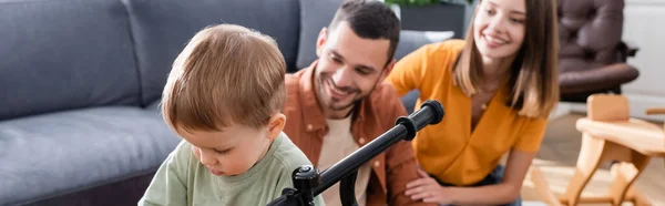 Toddler boy standing near bike and blurred parents at home, banner — Stock Photo