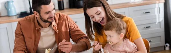 Jeune femme nourrissant son fils avec des pâtes près du mari à la maison, bannière — Photo de stock