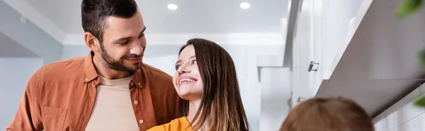 Smiling parents looking at each other near blurred kid in kitchen, banner — Stock Photo