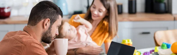 Jeune homme tenant une tasse près d'un ordinateur portable et une famille floue à la maison, bannière — Photo de stock