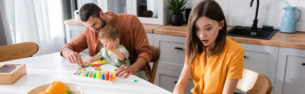 Shocked woman sitting near family playing educational game at home, banner — Stock Photo