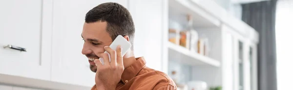 Cheerful man talking on mobile phone in kitchen, banner — Stock Photo