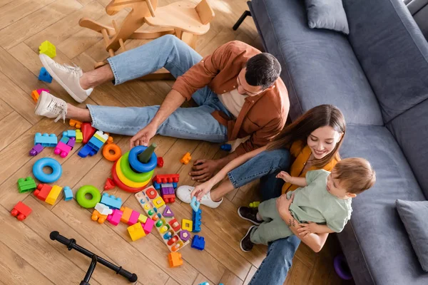 Vista ad alto angolo di madre sorridente che abbraccia il figlio vicino al marito e ai giochi a casa — Foto stock