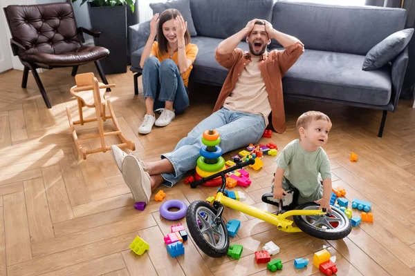 Kid standing near bike, building blocks and parents in living room — Stock Photo