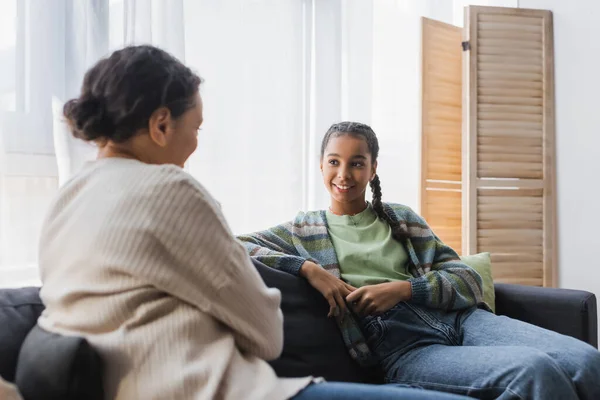 Sorrindo afro-americano adolescente menina falando com mãe turva no sofá em casa — Fotografia de Stock