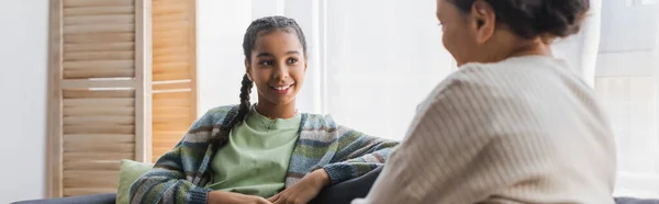 Lächelndes afrikanisch-amerikanisches Mädchen sieht verschwommene Mutter während eines Gesprächs zu Hause an, Banner — Stockfoto