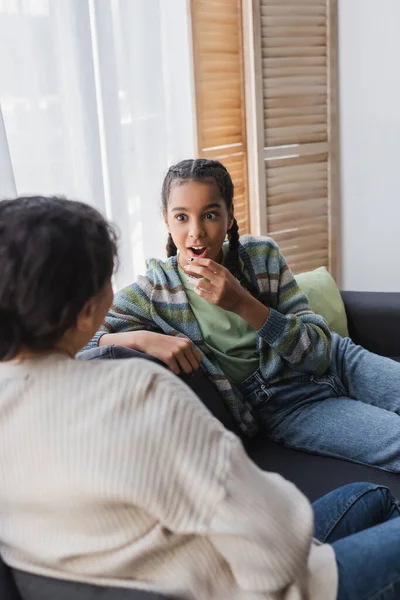 Amazed african american girl holding hand near face while listening to mom on couch at home — Stock Photo