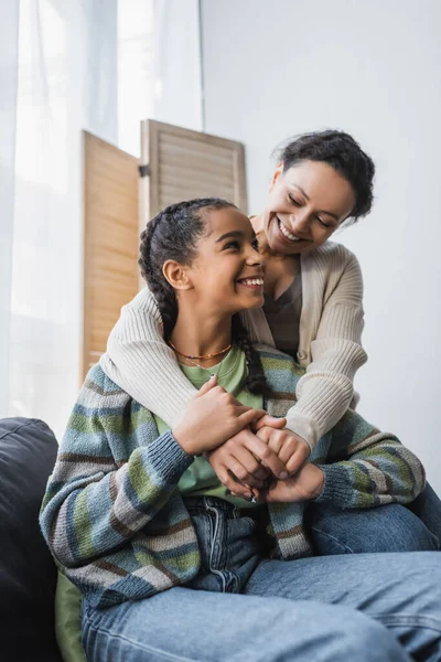 Feliz africana americana mujer abrazando adolescente hija sentado en sofá en casa - foto de stock