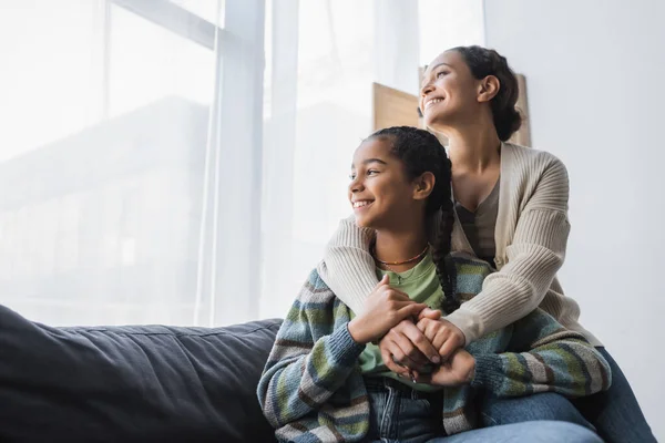 Happy african american mother and daughter sitting on couch and looking through window at home — Stock Photo