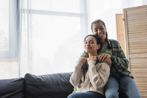 Adolescente afroamericana chica con los ojos cerrados abrazando mamá sentado en sofá cerca de ventana - foto de stock