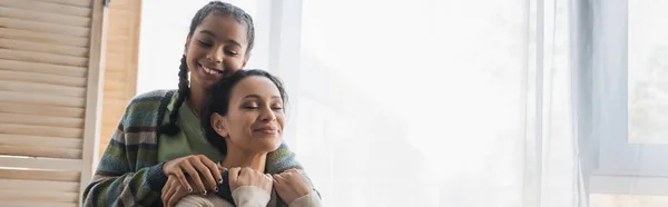 Adolescente afroamericano chica abrazando feliz mamá con los ojos cerrados cerca de ventana en casa, pancarta - foto de stock