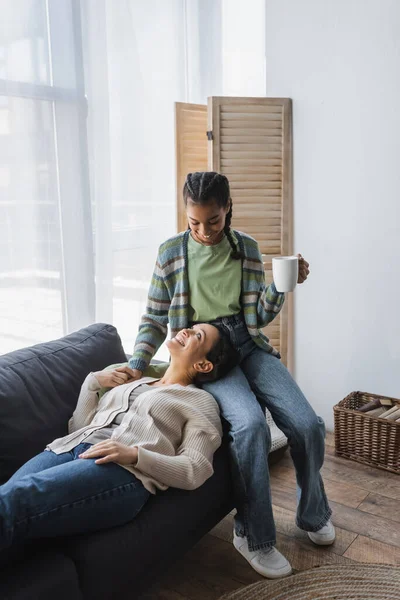 African american girl with cup of tea holding hands with happy mom lying on couch at home — Stock Photo