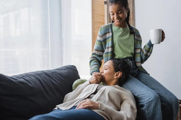 Mujer afroamericana acostada en un sofá y besándose la mano de una adolescente feliz sosteniendo una taza de té - foto de stock