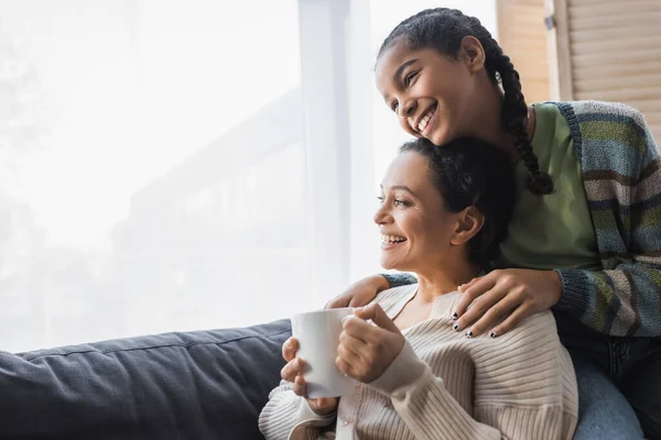 Fröhliche afrikanisch-amerikanische Teenager Mädchen umarmt glückliche Mutter sitzt auf der Couch mit Teetasse — Stockfoto