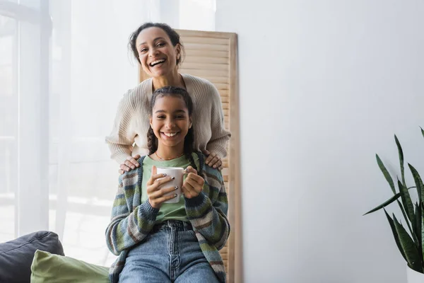 Alegre africano americano mujer y adolescente chica con taza de té sonriendo a cámara en casa - foto de stock