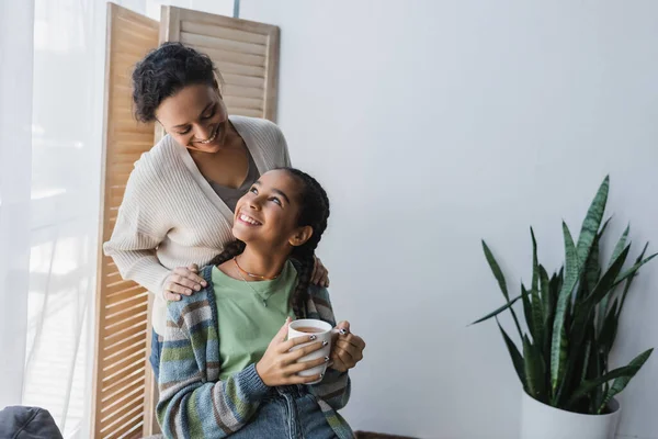 Feliz africano americano mujer tocando hombros de sonriente adolescente hija sosteniendo taza de té - foto de stock