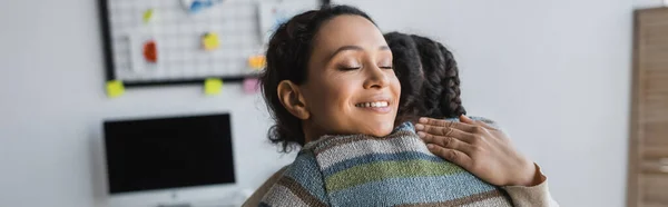 Cheerful african american woman with closed eyes hugging daughter at home, banner — Stock Photo