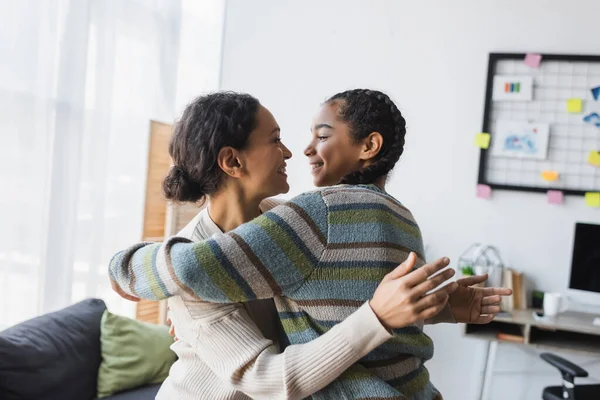 Freudige afrikanisch-amerikanische Mutter mit Teenie-Tochter, die einander ansieht, während sie sich zu Hause umarmt — Stockfoto