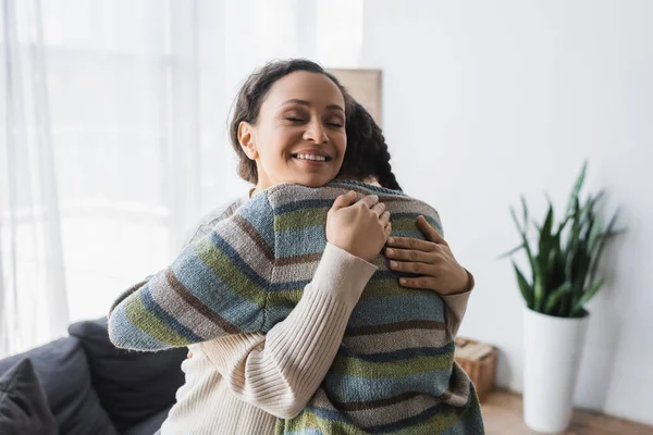 Gioiosa donna afroamericana con gli occhi chiusi abbracciando figlia adolescente a casa — Foto stock