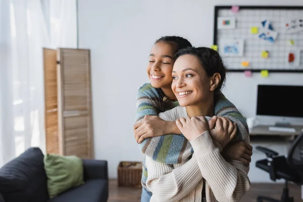 Smiling african american girl with closed eyes hugging happy mom near computer monitor on blurred background — Stock Photo