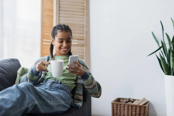 Sorridente ragazza adolescente africana americana che chatta sul telefono cellulare mentre tiene in mano una tazza di tè — Foto stock