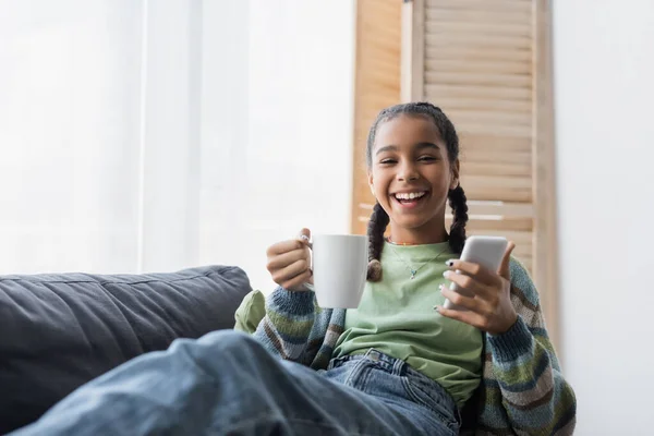 Cheerful african american teen girl looking at camera while holding smartphone and cup of tea — Stock Photo