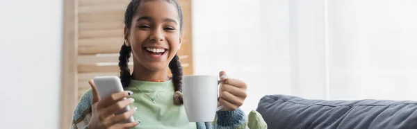 Joyful african american teenage kid with smartphone and tea cup smiling at camera, banner — Stock Photo