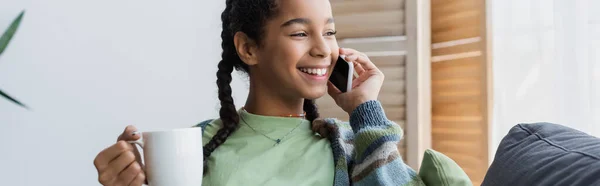 Chica afroamericana feliz sosteniendo taza de té durante la conversación en el teléfono celular, pancarta - foto de stock