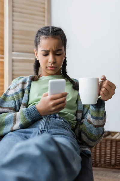 Displeased african american teenage girl with cup of tea messaging at cellphone at home — Stock Photo