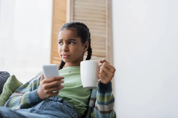 Upset african american teenager holding cup of tea and blurred mobile phone at home — Stock Photo