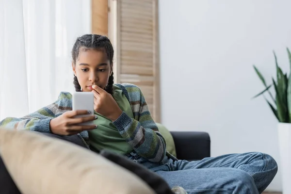 Pensive african american girl looking at mobile phone on sofa at home — Stock Photo