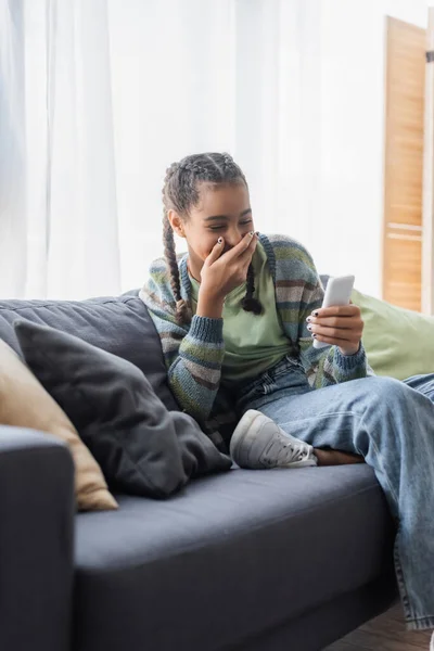 Africano americano ragazza ridere e copertura bocca con mano mentre seduto su divano e guardando smartphone — Foto stock