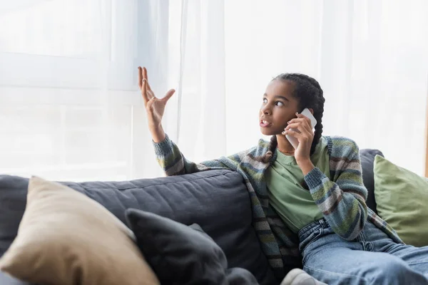 Adolescente afroamericana enojada haciendo gestos mientras habla por teléfono móvil en casa - foto de stock